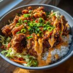 A close-up of a Bang Bang Chicken Bowl featuring crispy fried chicken, white rice, shredded slaw, and a creamy sauce, topped with green onions.