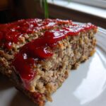 A close-up of a slice of Classic 1950s Meatloaf topped with a shiny ketchup glaze on a white plate.