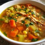 A close-up of a white bowl filled with Classic Caldo De Pollo Mexican Chicken Soup, featuring shredded chicken, carrots, celery, and corn, garnished with cilantro.