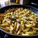 Close-up of Creamy Garlic Parmesan Chicken Pasta with penne pasta, chicken pieces, and parsley in a black pan.