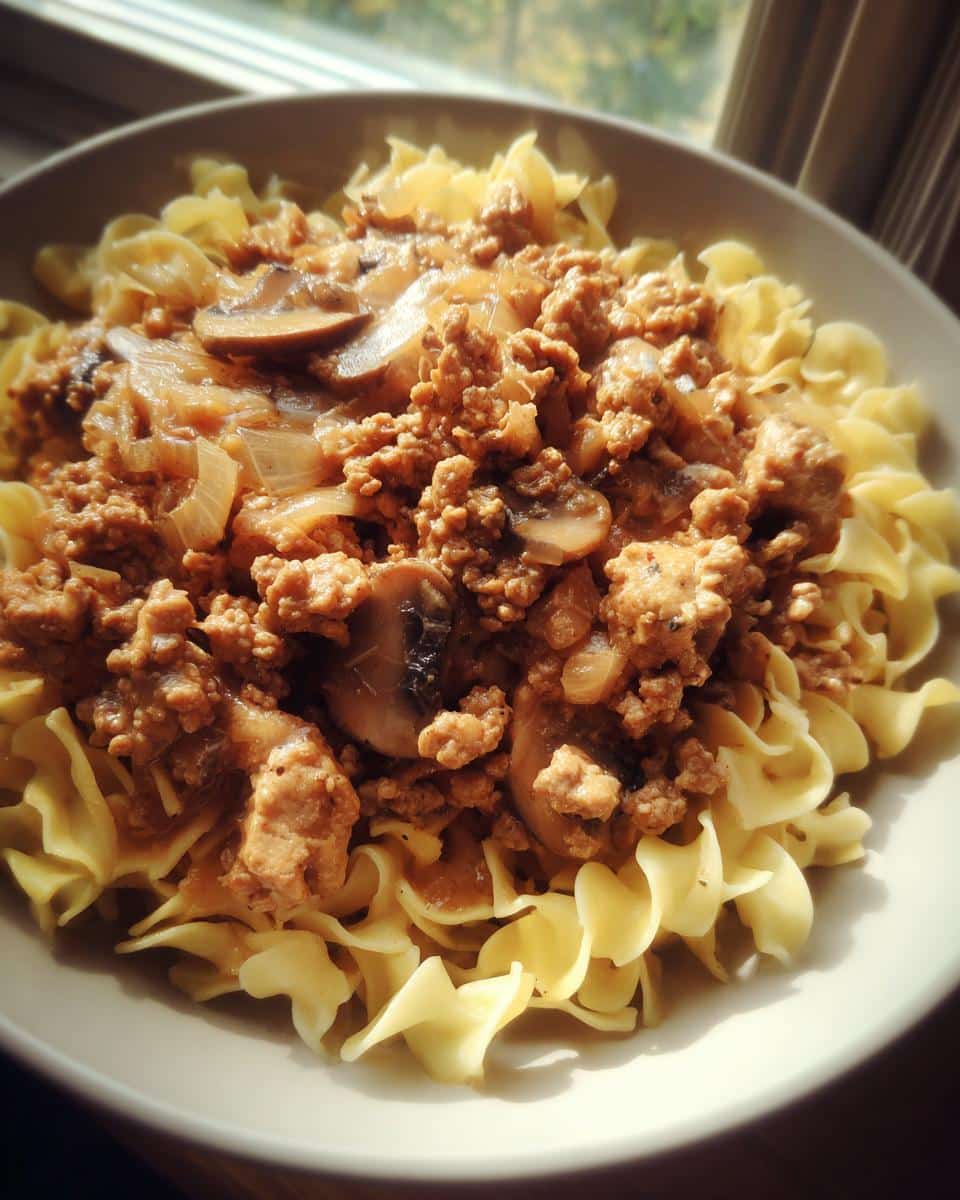 A close-up of a bowl filled with Easy Hamburger Stroganoff served over wide egg noodles, featuring ground beef, mushrooms, and onions.