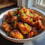 A bowl of glistening Easy Honey Sesame Chicken, coated in a sticky sauce and sprinkled with sesame seeds and green onions.