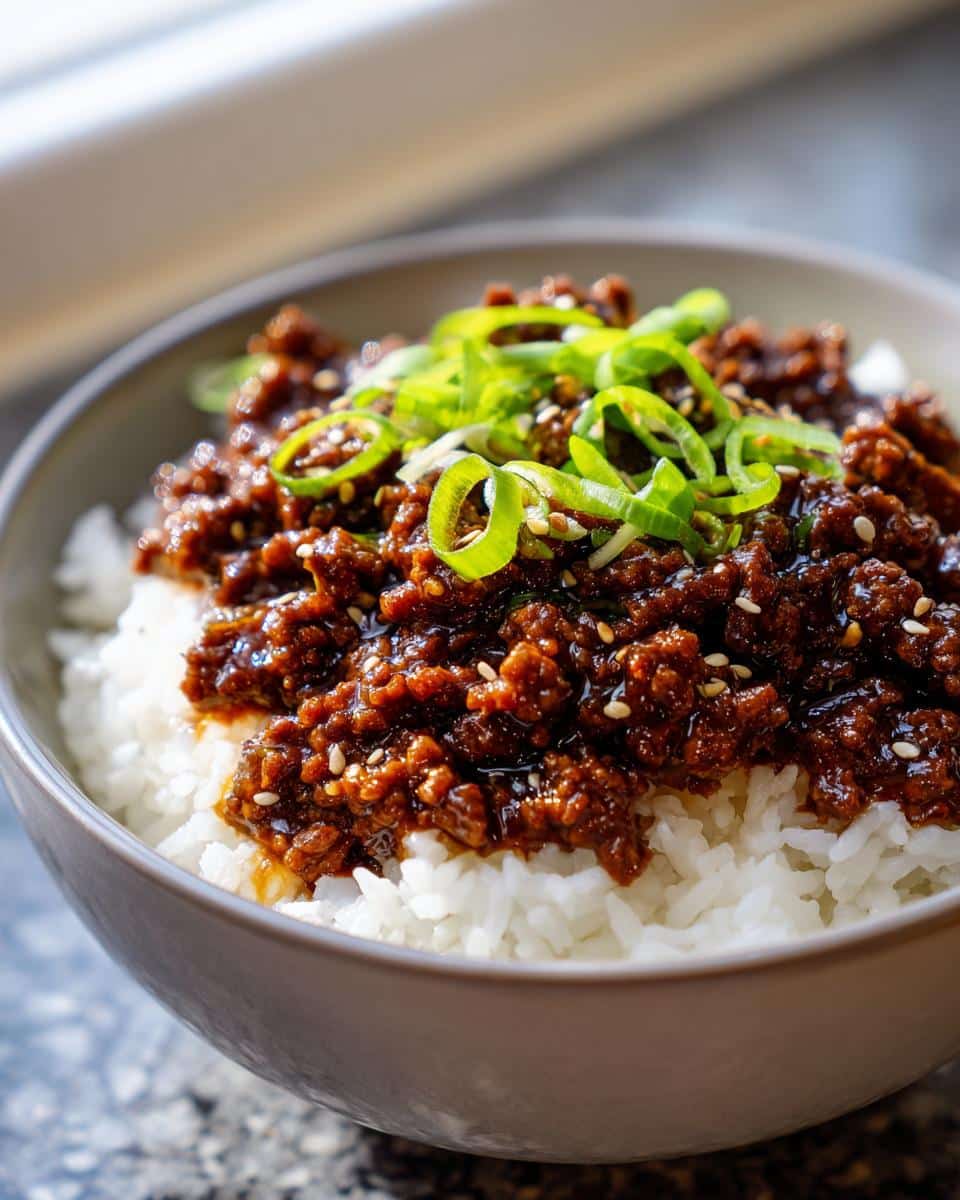 A bowl of white rice topped with savory Mongolian ground beef and garnished with green onions and sesame seeds.