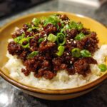 A bowl of fluffy white rice topped with glistening, saucy ground beef, garnished with chopped green onions and sesame seeds. This is an Easy Mongolian Ground Beef Recipe.