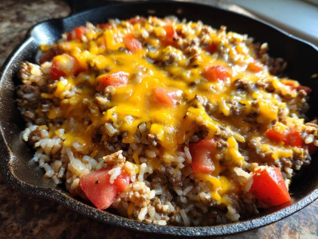 Close-up of a skillet filled with ground beef, rice, diced tomatoes, and melted cheddar cheese, a delicious ground beef recipe.