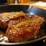 Close-up of two juicy Honey Mustard Air Fryer Pork Chops glistening in a pan with a honey mustard glaze and spices.