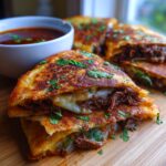 Close-up of crispy My Fave Birria Tacos filled with shredded beef and cheese, served with a side of consommé.