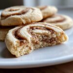 A close-up of a soft cinnamon roll sugar cookie with a bite taken out, revealing the swirled cinnamon filling.