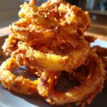A close-up, sunlit stack of golden-brown, crispy Spicy Cajun Onion Rings on a dark plate.