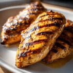 Close-up of three perfectly grilled chicken breasts with visible grill marks, glistening with marinade.