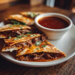 Close-up of crispy birria tacos filled with shredded beef and cheese, served with a side of rich birria tacos consome.