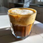 A close-up of a Cereal Milk Latte in a glass, topped with thick, creamy foam and rich espresso.