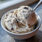 Two scoops of creamy cottage cheese ice cream with dark chocolate chips in a white bowl, with a spoon.