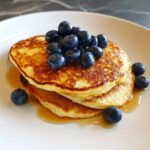 A stack of fluffy cottage cheese pancakes topped with fresh blueberries and drizzled with syrup.