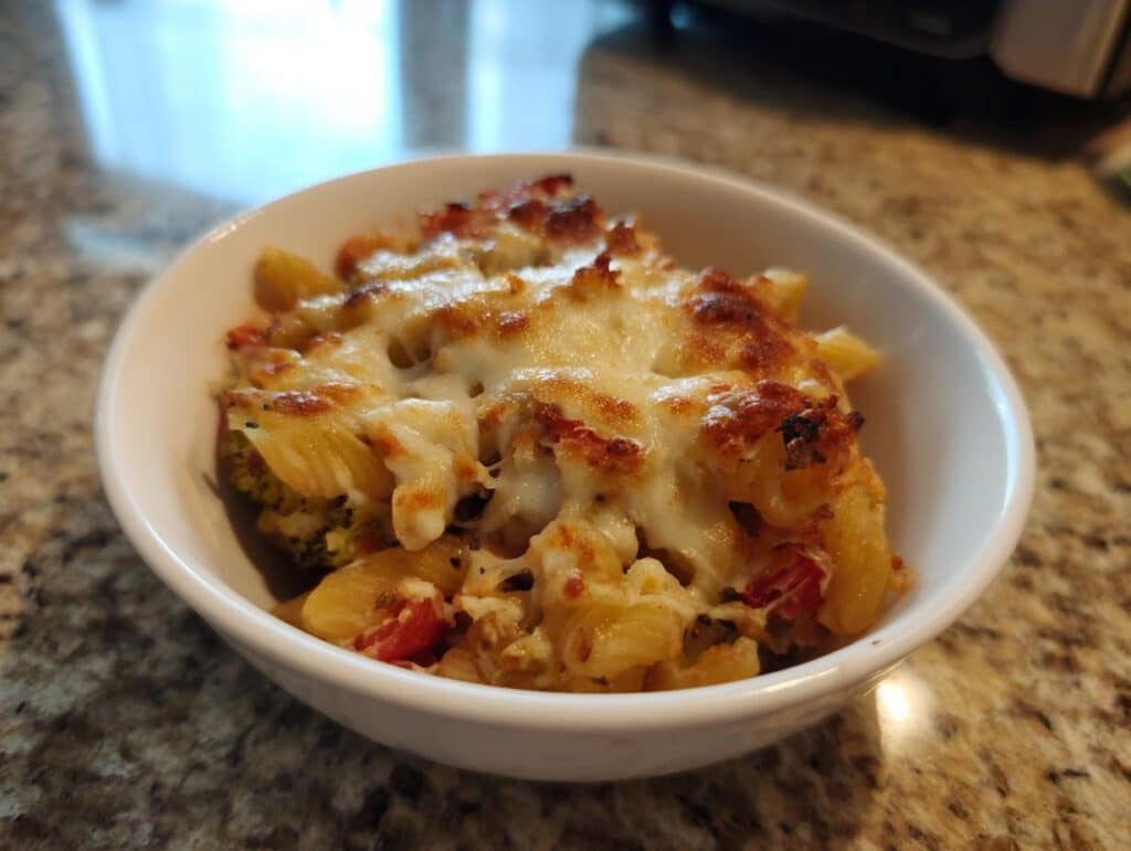 A close-up of a white bowl filled with a cheesy pasta bake, featuring penne pasta, broccoli, and tomatoes, perfect for family friendly pasta recipes.
