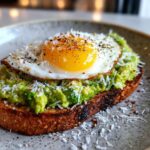 A close-up of Grated Egg Avocado Toast topped with a fried egg, shredded cheese, and spices.