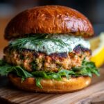 A close-up of a juicy grilled salmon burger topped with creamy dill sauce and fresh dill, served on a toasted bun with greens.