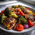 A plate of roasted chicken thighs with broccoli, cherry tomatoes, and red onion, seasoned with herbs. Perfect for healthy meal prep recipes.