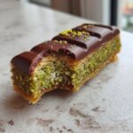 A close-up of a Knafeh Chocolate Bar (Dubai Chocolate) with a bite taken out, revealing layers of crispy pastry and pistachio filling.