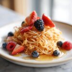 A close-up of Pancake Spaghetti topped with fresh strawberries, blueberries, raspberries, and blackberries, drizzled with syrup.