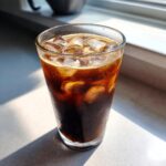 A tall glass filled with iced Proffee (Protein Coffee), showing dark coffee and ice cubes, with sunlight streaming in.