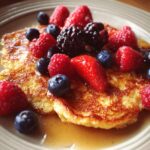 Close-up of golden-brown scrambled pancakes generously topped with fresh raspberries, blueberries, blackberries, and strawberries, drizzled with syrup.