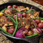 Close-up of a cast iron skillet filled with simple one pot meal: roasted chicken thighs, red potatoes, green beans, and red onions.