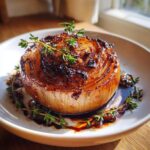 A perfectly cooked whole roasted onion, glazed and topped with fresh thyme sprigs, served in a white bowl with a rich sauce.