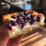A hand holding a slice of Blueberry Cream Cheese Bars, showcasing a creamy filling and fresh blueberries.