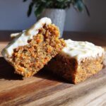 Two frosted Carrot Cake Bars, one cut open to show the texture, sitting on a wooden board.