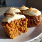Four carrot cake cupcakes with cream cheese frosting. One cupcake is cut in half to show the texture.
