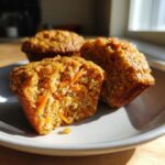 Three carrot oat muffins on a plate, one muffin is halved to show the texture and shredded carrots inside.