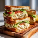 A tall, stacked Chicken Avocado Sandwich with tomato and lettuce on toasted bread, sitting on a wooden board.