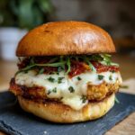 Close-up of a Chicken Parmesan Burger with melted cheese, sun-dried tomatoes, and arugula on a brioche bun.