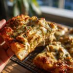 A hand holds a slice of Chicken Pesto Pizza with melted cheese and herbs, showcasing the delicious layers.