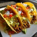 Three chicken soft tacos filled with shredded chicken, lettuce, cheese, tomatoes, and sour cream on a white plate.