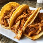 Three mouthwatering chicken tacos crockpot filled with shredded seasoned chicken on a white plate.