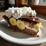 A delicious slice of Chocolate Banana Cream Pie with whipped cream, bananas, and chocolate topping.