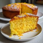 A slice of Citrus Olive Oil Cake on a plate, garnished with orange zest and powdered sugar.
