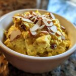 A bowl of Coronation chicken salad, topped with sliced almonds, presented on a granite countertop.