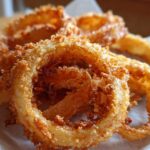 Close-up of golden, crispy onion chips. The onion rings are piled on a white surface, showcasing their texture.