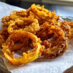 A pile of golden, deep-fried crispy onion chips on a white paper towel, ready to eat.