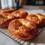 Close-up of freshly baked homemade bagels recipe, generously topped with golden sesame seeds, resting on a cooling rack.
