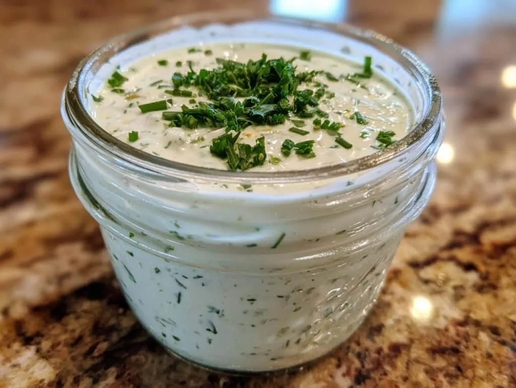 A close-up of a glass jar filled with creamy homemade ranch dressing, garnished with fresh chives and parsley.