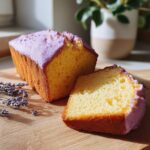 A slice of moist yellow lavender cake with a light purple icing, served on a wooden board with dried lavender sprigs.