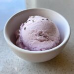 A close-up of a scoop of pale purple lavender ice cream in a white bowl, garnished with dried lavender buds.