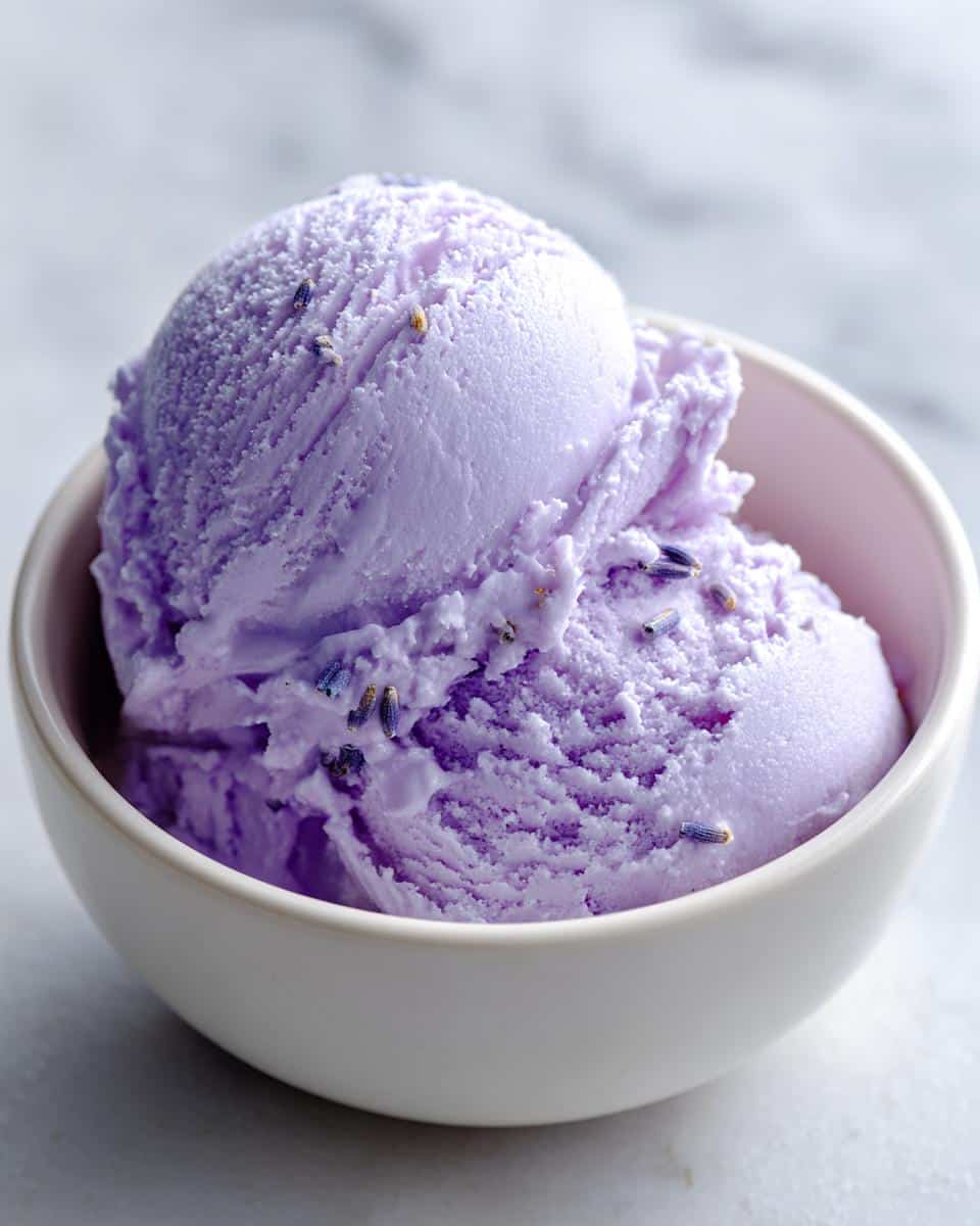 Two scoops of creamy purple lavender ice cream in a white bowl, garnished with dried lavender buds.
