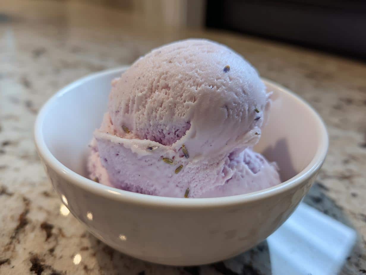A close-up of two scoops of light purple lavender ice cream in a white bowl, with visible lavender buds.