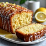 A close-up of a Lemon Drizzle Cake with a slice cut, drizzled with icing and lemon zest.