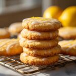 A stack of four glazed Lemon Shortbread Cookies on a wire rack, with lemons in the background.